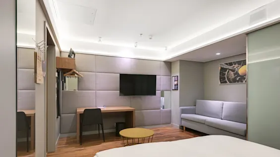 En dubbelsäng ligger i ett modernt hotellrum med en väggmonterad TV, ett skrivbord av trä och en grå soffa, vilket skapar en ren och bekväm miljö.