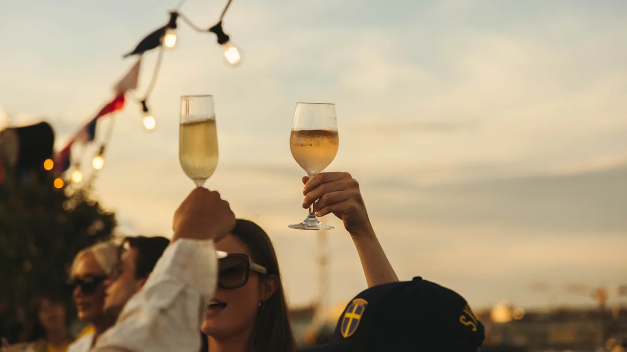 Folk skåler med drinker på et utendørs arrangement under lyslenker, feirer på Stockholm Under Stjärnorna.