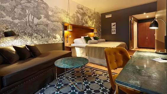 A tastefully decorated hotel room with a bed, a wall with a forest mural, and stylish furniture.