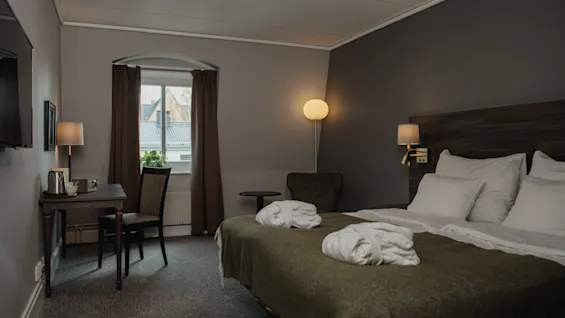 En säng prydd med vita morgonrockar i ett varmt belyst hotellrum, med skrivbord, stol, fönster med gardiner och golvlampa i en mysig miljö.