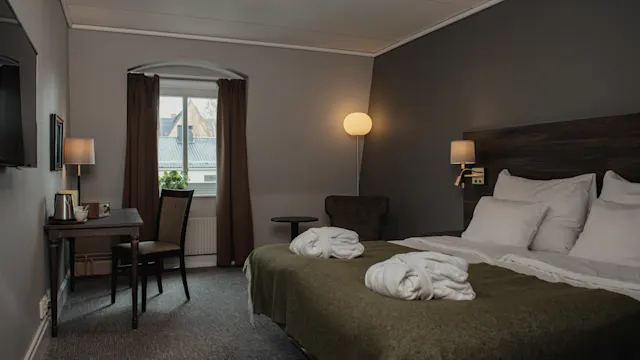 A bed adorned with white robes rests in a warmly lit hotel room, featuring a desk, chair, window with curtains, and a floor lamp in a cozy atmosphere.