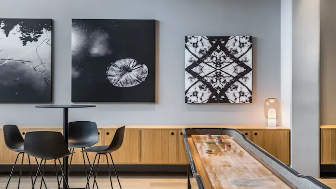 A table with four black chairs sits in a modern room. Two abstract black-and-white artworks hang on the grey wall, beside a wooden shuffleboard and a lamp.
