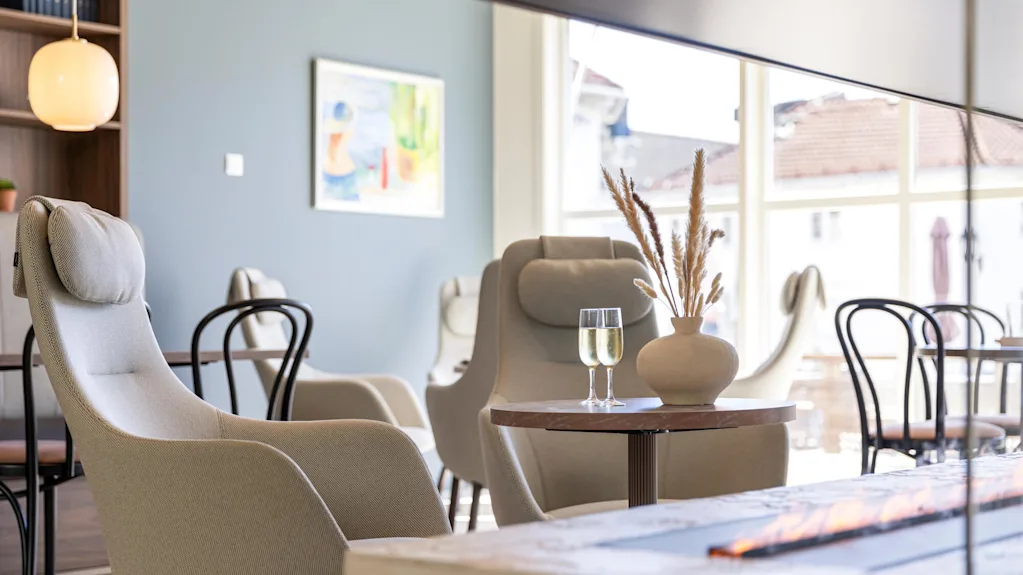 Moderne møbler i et veloplyst rum med store vinduer og kunst på væggen; vandglas på et bord ved siden af en komfortabel stol.