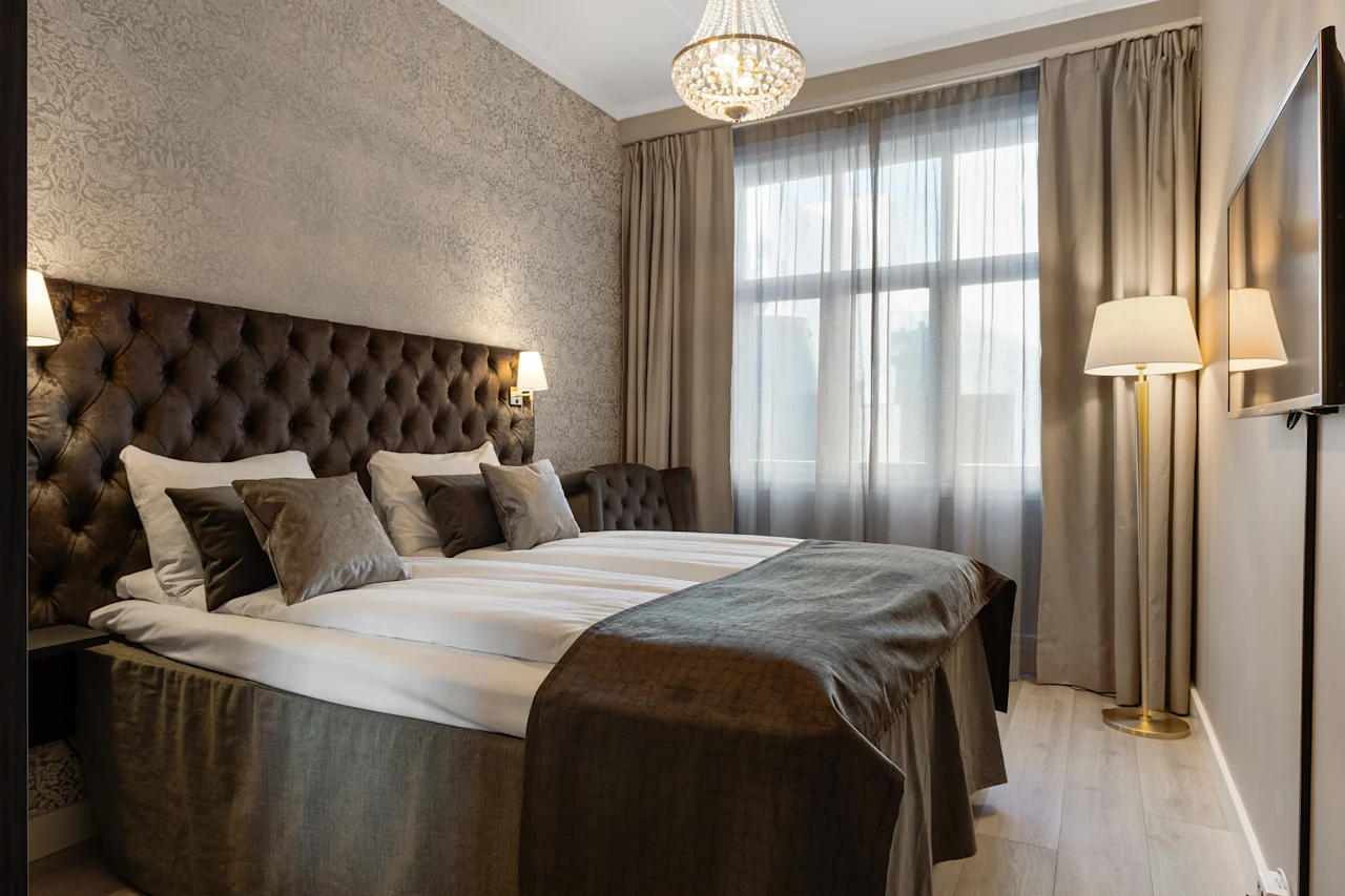 Et komfortabelt og elegant hotelværelse med en stor seng, tuftet sengegavl, lysekrone og vindue med gardiner.