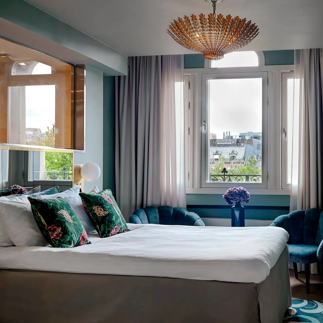 A bed with floral cushions stands in front of a window, flanked by blue chairs, beneath an elegant chandelier in a stylishly decorated bedroom.