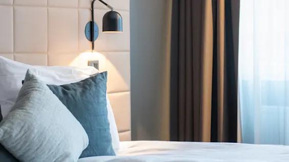 A neatly made bed with white linens and blue pillows sits against a cushioned headboard. A wall-mounted lamp and dark curtains frame a softly lit window nearby.