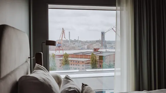 En säng med flera kuddar i ett hotellrum står i förgrunden och har utsikt över ett stort fönster som visar industriella kranar och byggnader utanför under en molnig himmel.