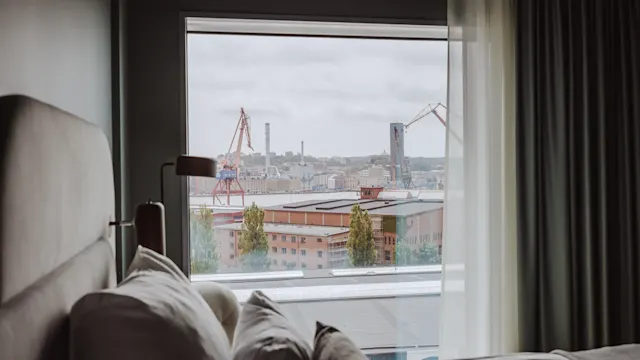 En seng med flere puder i et hotelværelse står i forgrunden og giver udsigt til et stort vindue, der viser industrikraner og bygninger udenfor under en overskyet himmel.