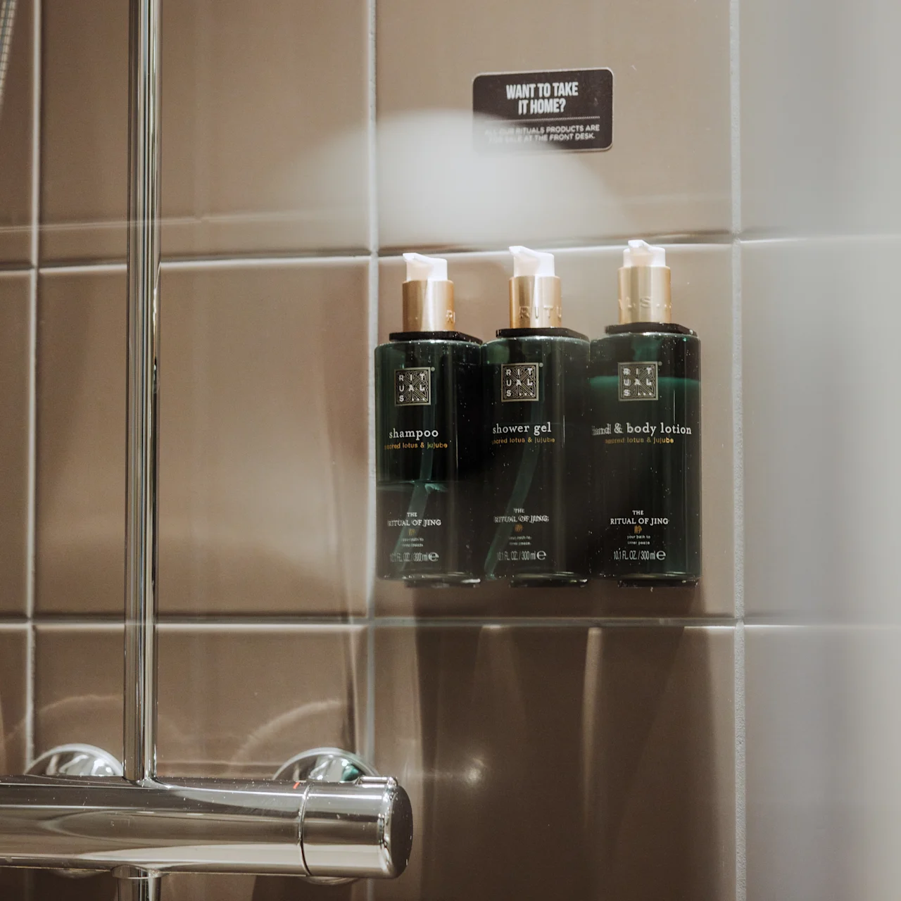 Schampo, duschgel och bodylotionflaskor är prydligt placerade på en hylla i ett badrum mot bruna kakelväggar. Texten lyder: "Want to take it home? If so, we charge you 12,50 EUR."