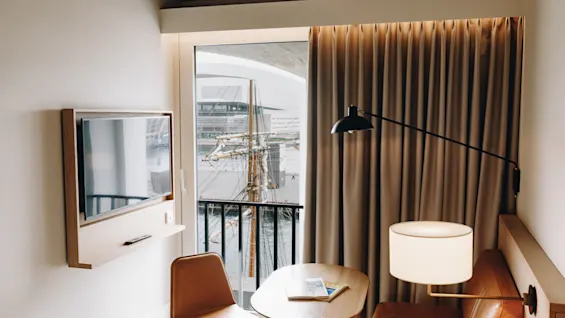 Ett mysigt rum med utsikt över en hamn, med en TV, ett litet bord, en stol och en bekväm bänk med en lampa.