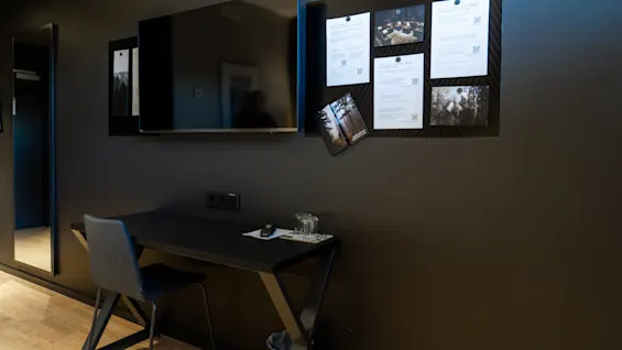 En väggmonterad TV ovanför ett skrivbord med en stol, bredvid en anslagstavla med informationspapper och fotografier i ett rum med trägolv. Text inkluderar "MOVEMENT," "FÖRSÖ PARK SPA," "BEHANDLINGAR."