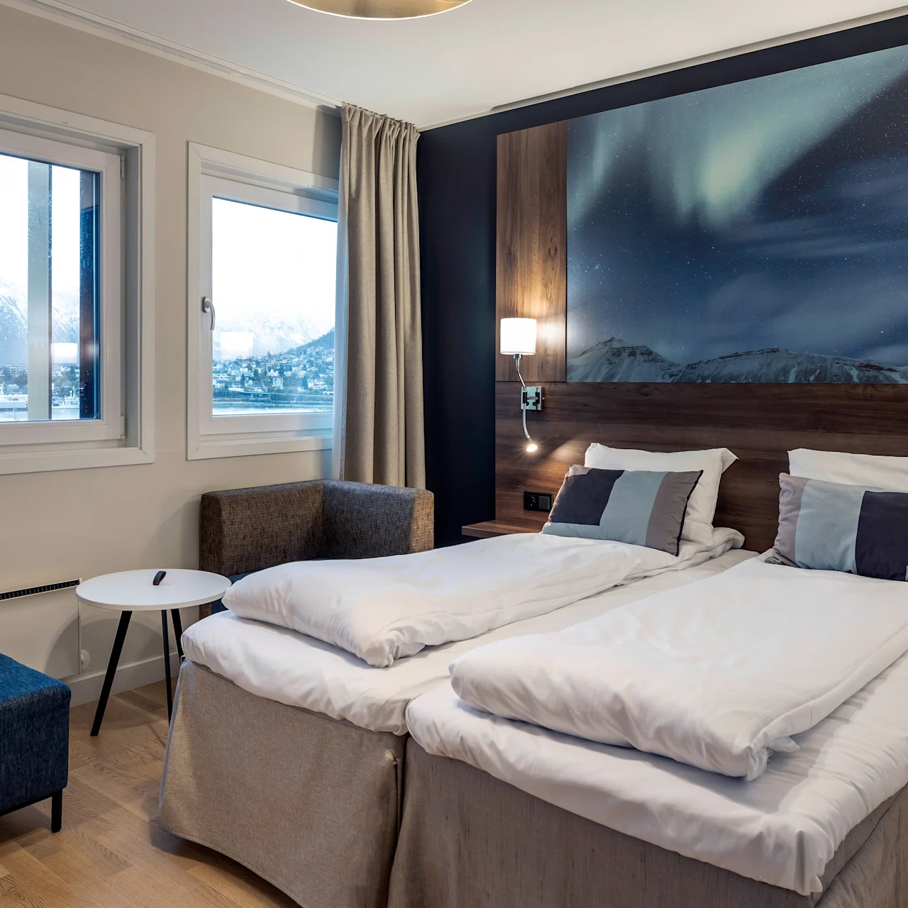 A neatly made double bed with white linens, framed by bedside lamps and an aurora-themed artwork. Chairs and a small table by the window overlook snowy mountains.