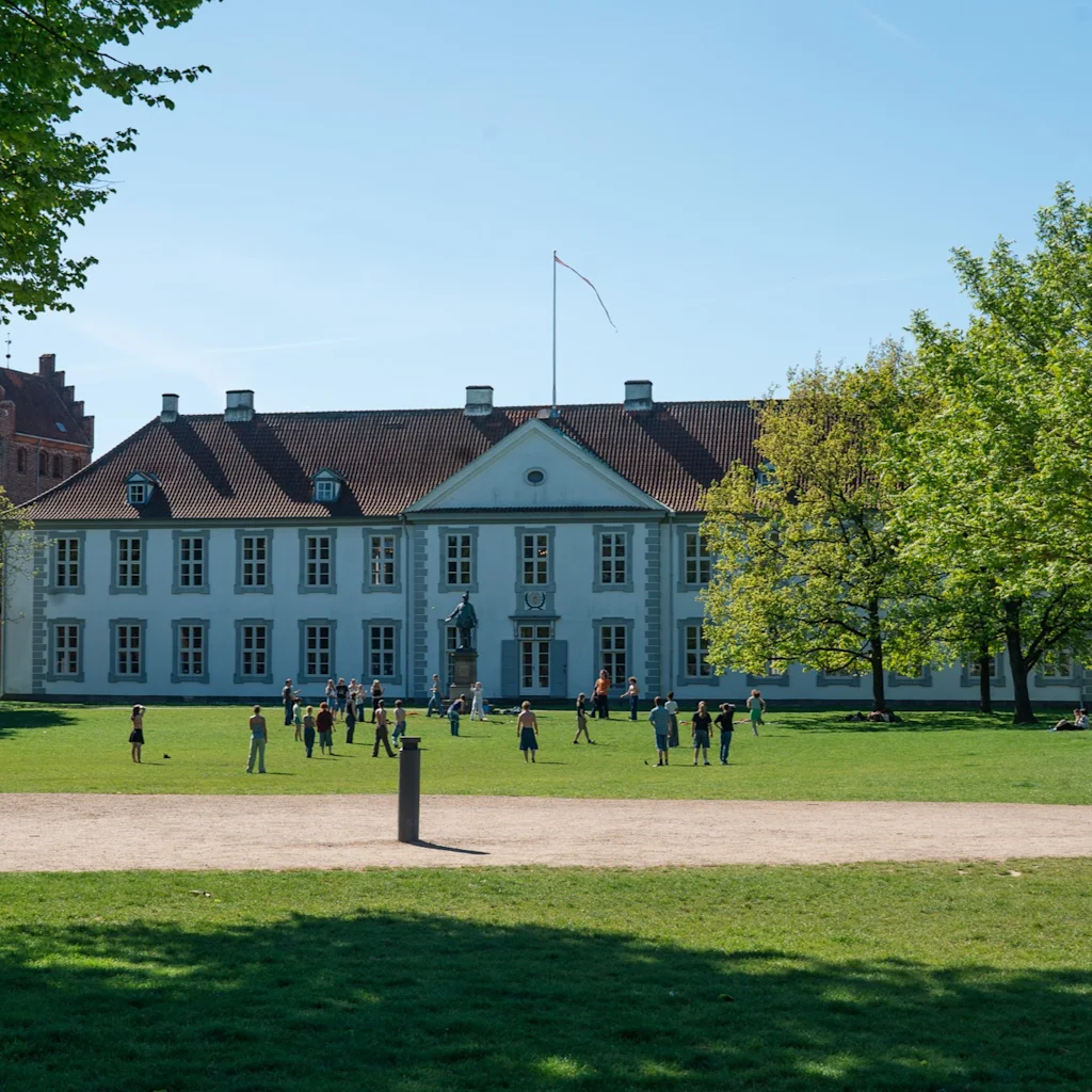 Människor njuter av en solig dag på den gröna gräsmattan framför Odense slott och Kungens trädgård, med träd som omger området.