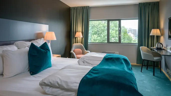 A neatly made bed with teal accents sits in a softly lit hotel room, flanked by a chair and desk. Large window shows greenery outside.