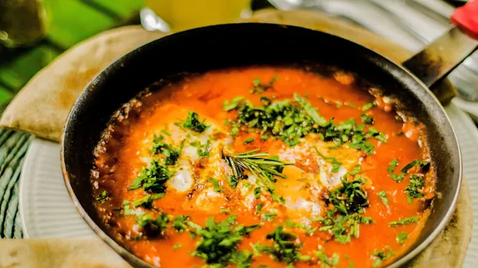 A skillet with tomato-based dish garnished with herbs beside flatbread on a table.
