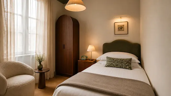 Ett varmt och inbjudande hotellrum med en enkelsäng, fåtölj och garderob. Naturligt ljus. Våra rum tar fortfarande form.
