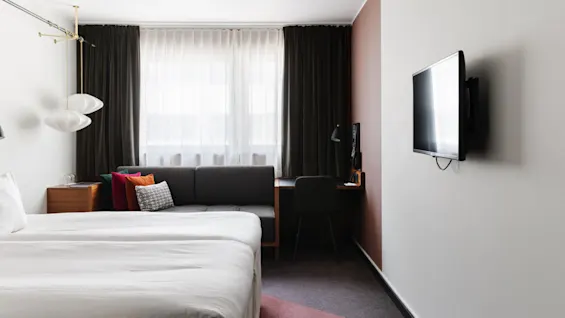 Ett snyggt bäddad säng i ett modernt hotellrum med soffa, skrivbord och väggmonterad TV.
