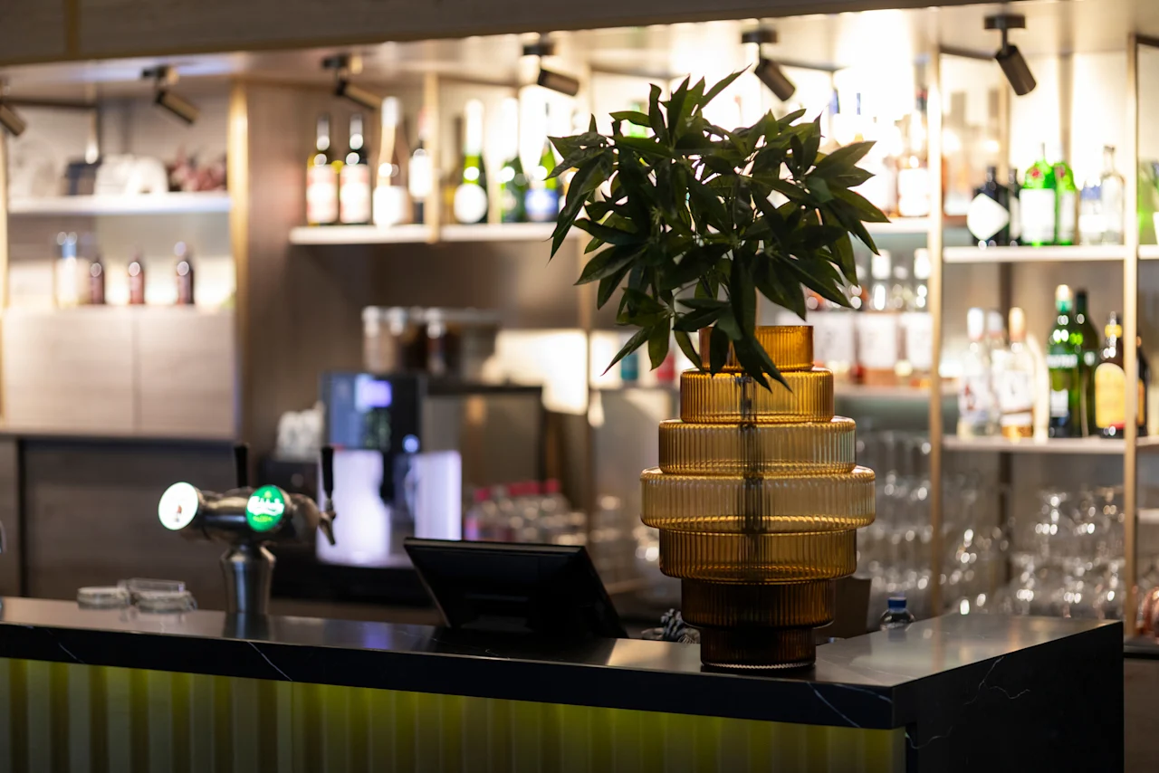 A stylish bar counter at Home Hotel Grand Bodø with a beer tap, a plant in a decorative vase, and shelves of bottles in the background.