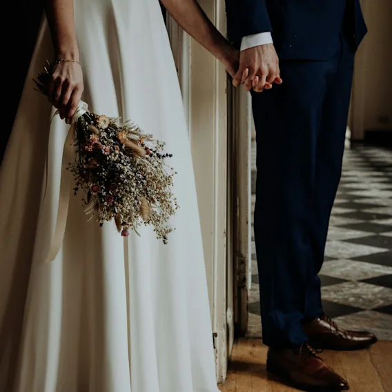 A couple holding hands, one in a white dress with a bouquet, the other in a suit, standing in a hallway with a checkered floor.