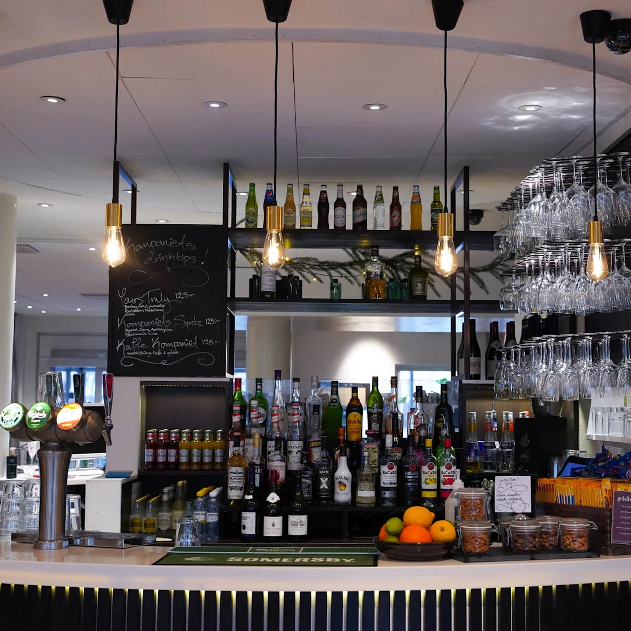 An empty bar, with assorted drinks displayed, and pendant lights hanging above in an indoor setting. Text on board: "Champagne & cocktails", "Your choice 125", "Non-alcoholic Spritz 125", "Kopparbergs Non-alcoholic pear 125", "Ginger beer".