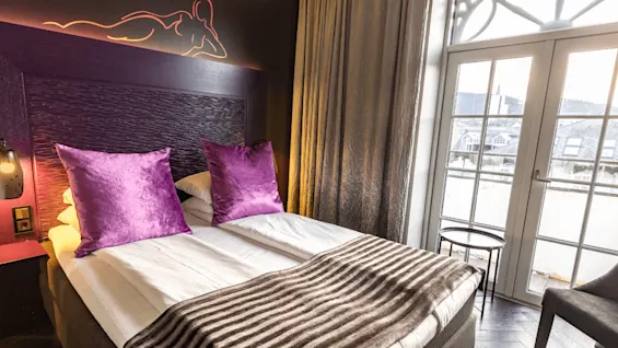 A bed with purple pillows and a striped blanket is under a decorative headboard. The setting includes a wall lamp, large curtains, and a window overlooking a cityscape.