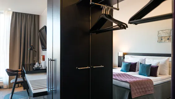 En garderobe med hengende klær deler et hotellrom med en pent oppredd seng, skrivebord med lampe og veggmontert TV. Myke lys og gardiner skaper en koselig atmosfære.