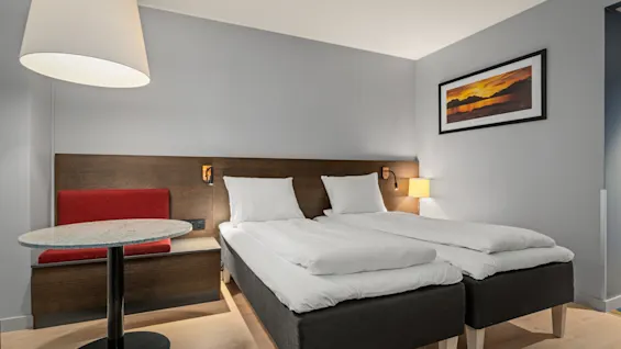 Et hotelværelse har to pænt opredte senge, ledsaget af et lille bord og en stol. Værelsets vægge er lyse, udsmykket med landskabskunst og blidt oplyst af en lampe.