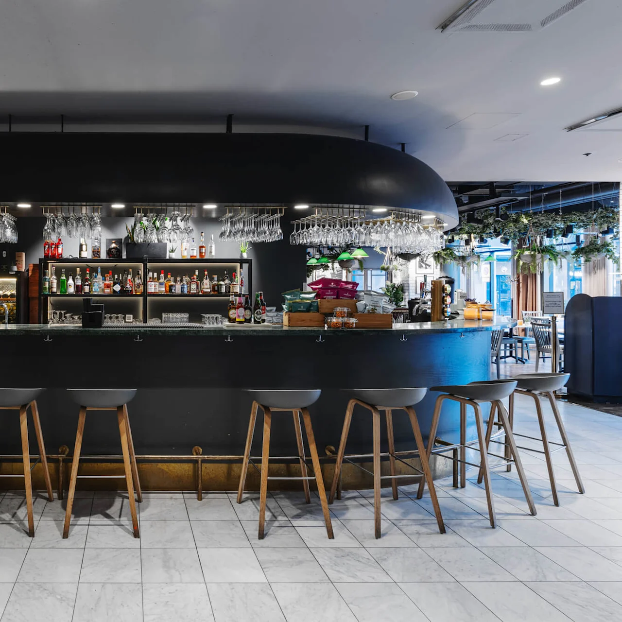A modern bar with a black countertop features various bottles and glassware. Wooden stools line the bar, surrounded by sleek interior decor and overhead lighting.