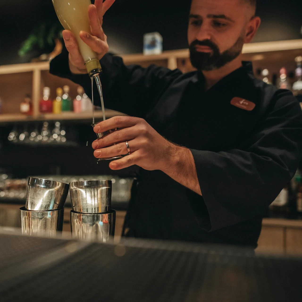 En bartender heller væske fra en flaske i en metallshaker på en bar.