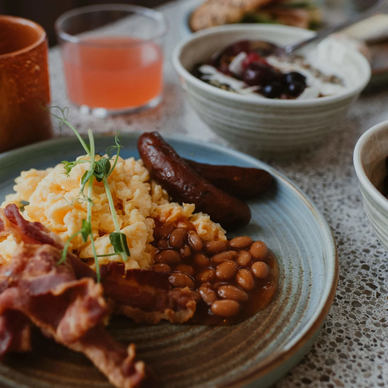 A plate with scrambled eggs, sausages, bacon, and baked beans, is accompanied by a glass of pink juice, a bowl of yogurt with berries, and bread in a breakfast setting.