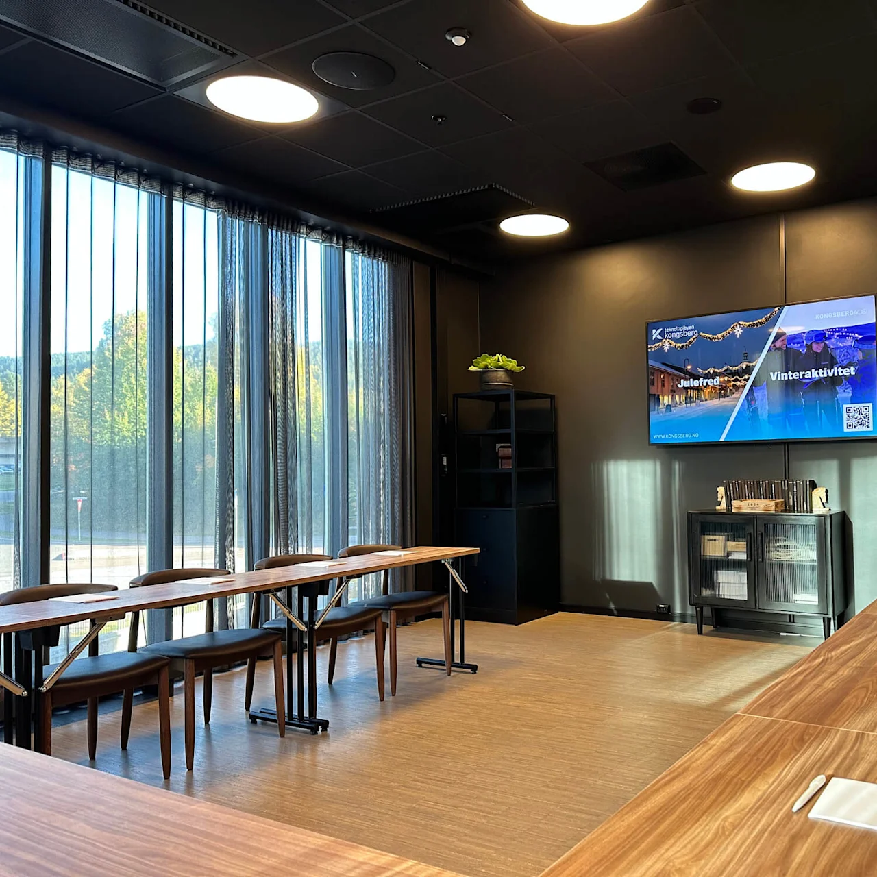 An office break room with a large TV on the wall displaying advertising messages, wooden tables, and chairs set for eating, located next to large windows.