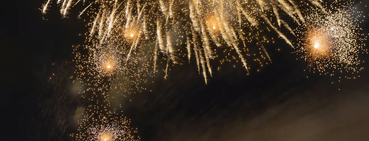 Fireworks explode in a night sky, illuminating the dark backdrop with golden light. A crowd of onlookers observes the spectacle, capturing the moment with cameras and phones.