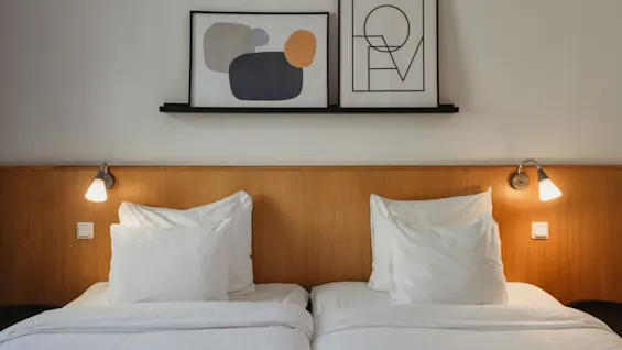 Kaksi mukavaa sänkyä valkoisilla liinavaatteilla ja puisella sängynpäädyllä, koristeltu modernilla taiteella, Home Hotel Uppsalassa.