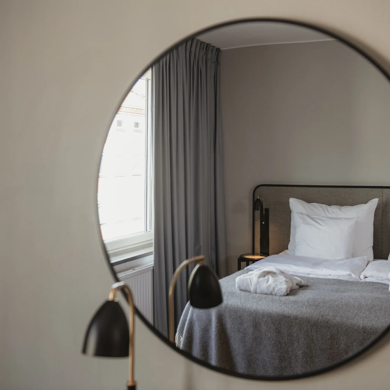 A round mirror reflects a neatly made bed with white pillows and a gray blanket, next to a window with curtains in a softly lit room.