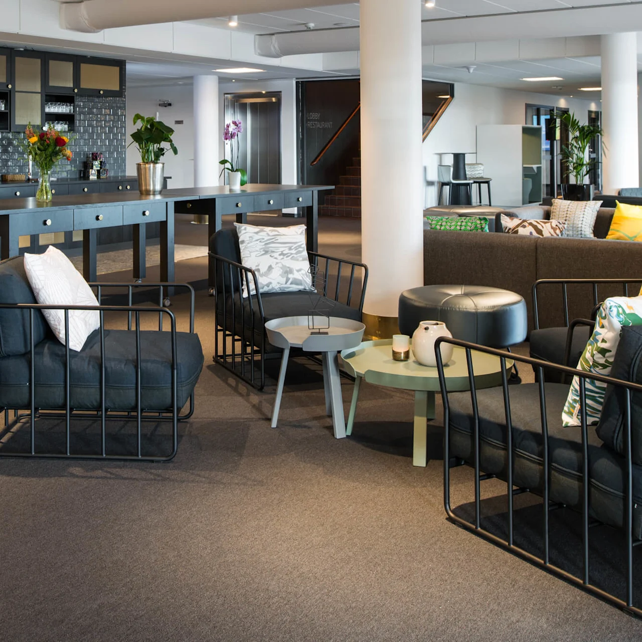 Fåtöljer står i ett modernt loungeområde, omgivet av bord och soffor, med växter och samtida inredning. En mörk kaklad disk med blommor i bakgrunden. Skyltar säger "Lobby Restaurant."