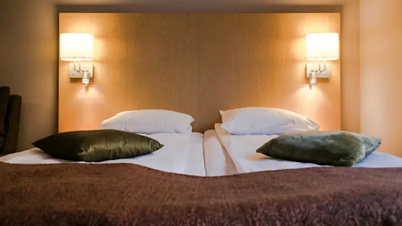 A neatly made double bed with two pillows and green cushions is flanked by wall-mounted lamps on a wooden headboard, set in a softly lit bedroom.