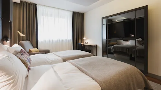 To omhyggeligt redte enkeltsenge står i et moderne hotelværelse, med en grå lænestol og skrivebord foran store draperede vinduer, reflekteret i et vægspejl.