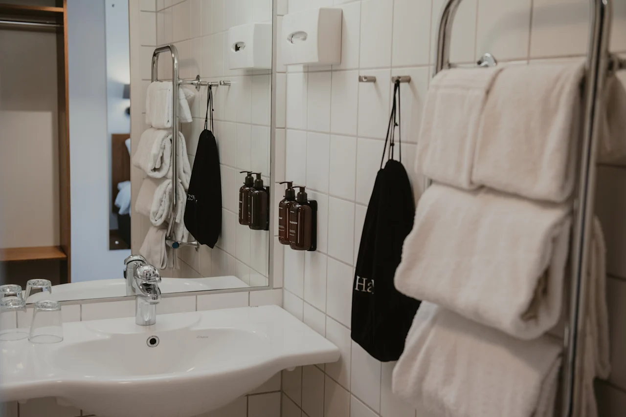 Siisti ja moderni hotellin kylpyhuone, jossa on pesuallas, peili, puhtaita valkoisia pyyhkeitä ja hygieniatuotteita. Musta pyyhe, jossa 'Ha', näkyy.