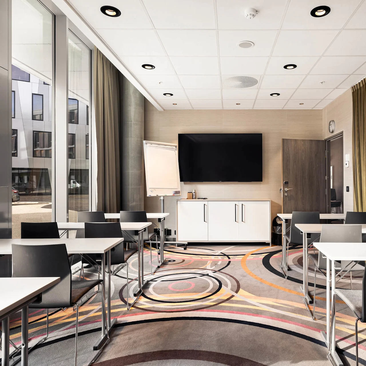 Tables and chairs are arranged in a modern conference room. A large TV and flip chart stand against the wall. Sunlight streams through large windows, illuminating the space.