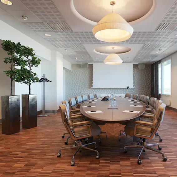 Ett långt konferensbord är omgivet av stolar på hjul, utrustat med anteckningsblock och vattenflaskor. Rummet har stora fönster, moderna taklampor och dekorativa väggpaneler.