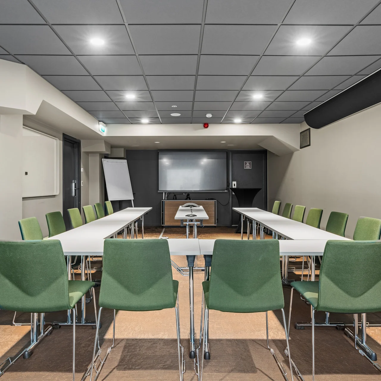 Et konferencelokale med U-formet borde; grønne stole placeret rundt. Et whiteboard og flipchart står foran. Lys belysning på et mørkt loft skaber en moderne setting.