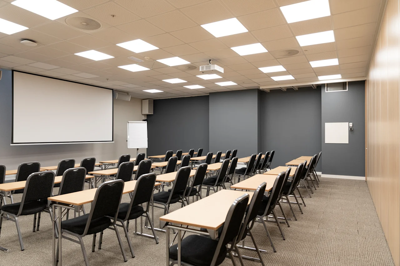 A modern conference room with rows of tables and chairs facing a large projection screen and a whiteboard.