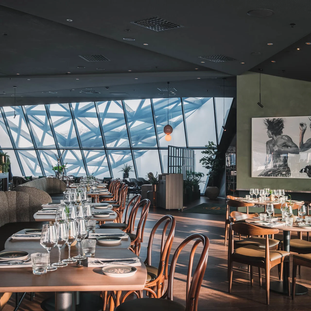 Luxurious restaurant with neatly set tables and chairs, featuring glassware and napkins. Sunlight streams through large geometric windows, creating a modern, airy atmosphere. Artworks adorn the walls, enhancing elegance.