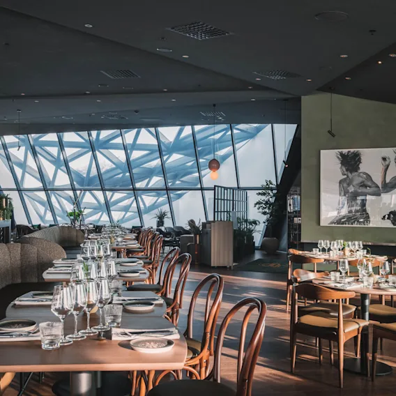 Luksuriøs restaurant med pænt dækkede borde og stole, med glas og servietter. Sollys strømmer gennem store geometriske vinduer, hvilket skaber en moderne, luftig atmosfære. Kunstværker pryder væggene og fremhæver elegancen.