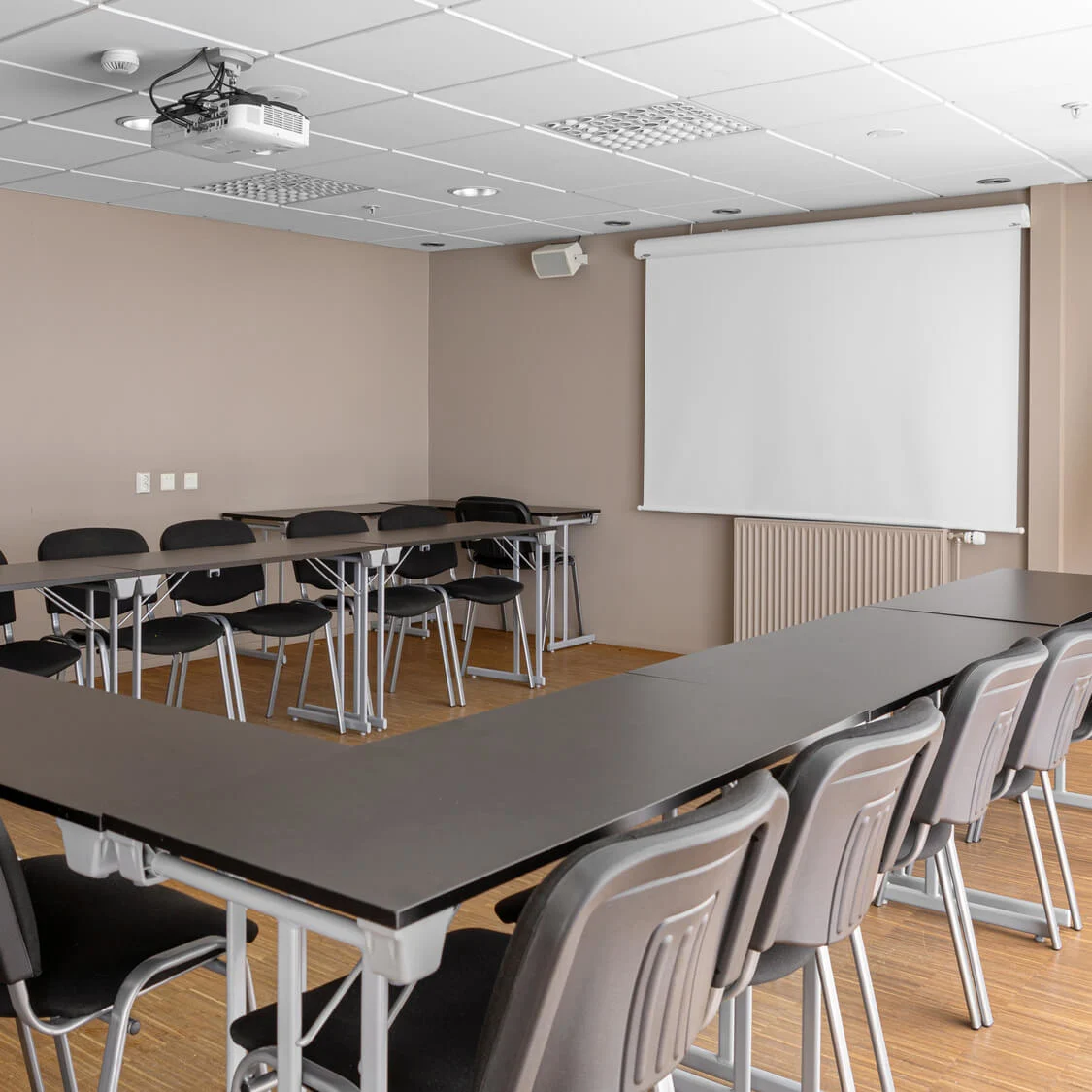 Ett U-format konferensbord med svarta stolar fyller ett rum med en projektor i taket, en vit projektionsskärm på väggen och ett blädderblocksställ i närheten.