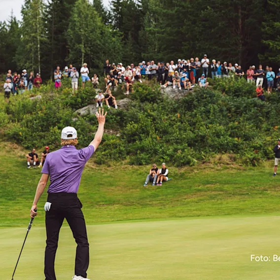 En golfspiller sett bakfra, kledd i lilla skjorte og hvit caps, som holder en golfkølle og vinker til et stort publikum samlet på en gresskledd bakke på en golfbane.