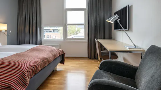 En pænt redt seng med stribet tæppe ved siden af et vindue, ved siden af et skrivebord med en lampe. Stol og TV fuldender det moderne rum. Tekst på bygningen udenfor: "TORDEL OFNUNG."