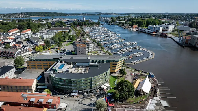 A curved, modern building with green and yellow panels overlooks a marina filled with docked boats. Surrounding the area are residential buildings and a vibrant, green landscape stretching to the horizon.