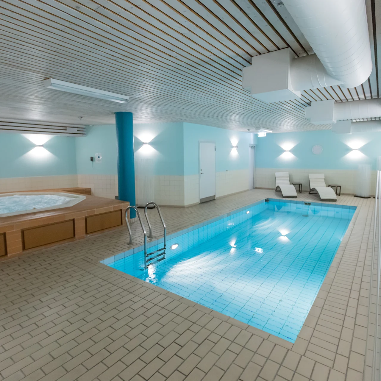 Pool with clear water and adjacent hot tub in a tiled, well-lit indoor space. Lounge chairs are positioned on the perimeter for relaxation, under a wooden slat ceiling.