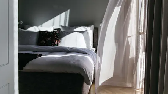 En pent seng med hvite laken og pynteputer står i et solfylt rom. Gjennomsiktige gardiner vaier forsiktig i sollyset fra et åpent vindu.